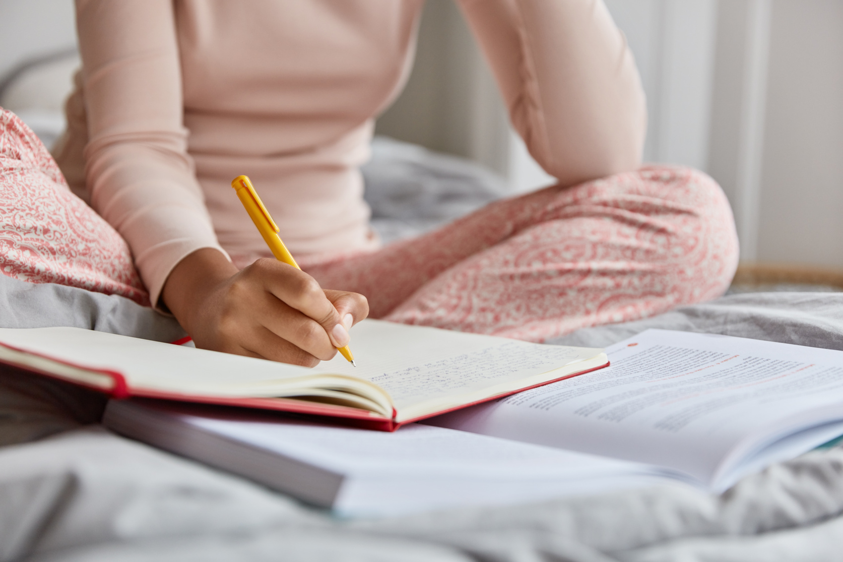 Cropped image of unrecognizable woman in nightclothes, writes down information in notepad, rewrites topic from textbook, poses at bed alone, has nice handwriting. Close up shot, focus on writing